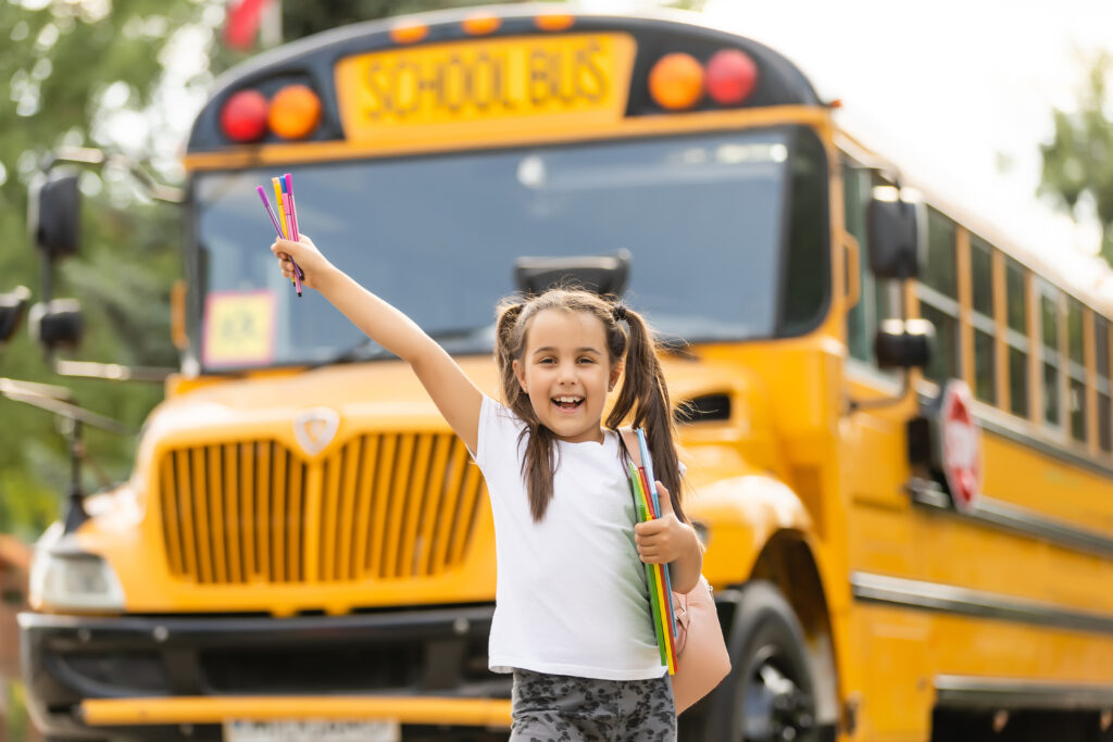 Smiling young student holding school supplies and raising hand in excitement in front of a yellow school bus.